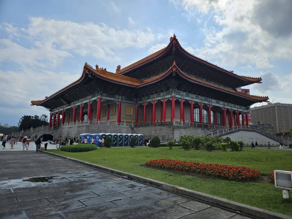A grand traditional Chinese-style building in Taipei, featuring a sweeping orange-tiled roof, red columns, and intricate architectural details. The structure is set against a partly cloudy sky with manicured greenery in the foreground.