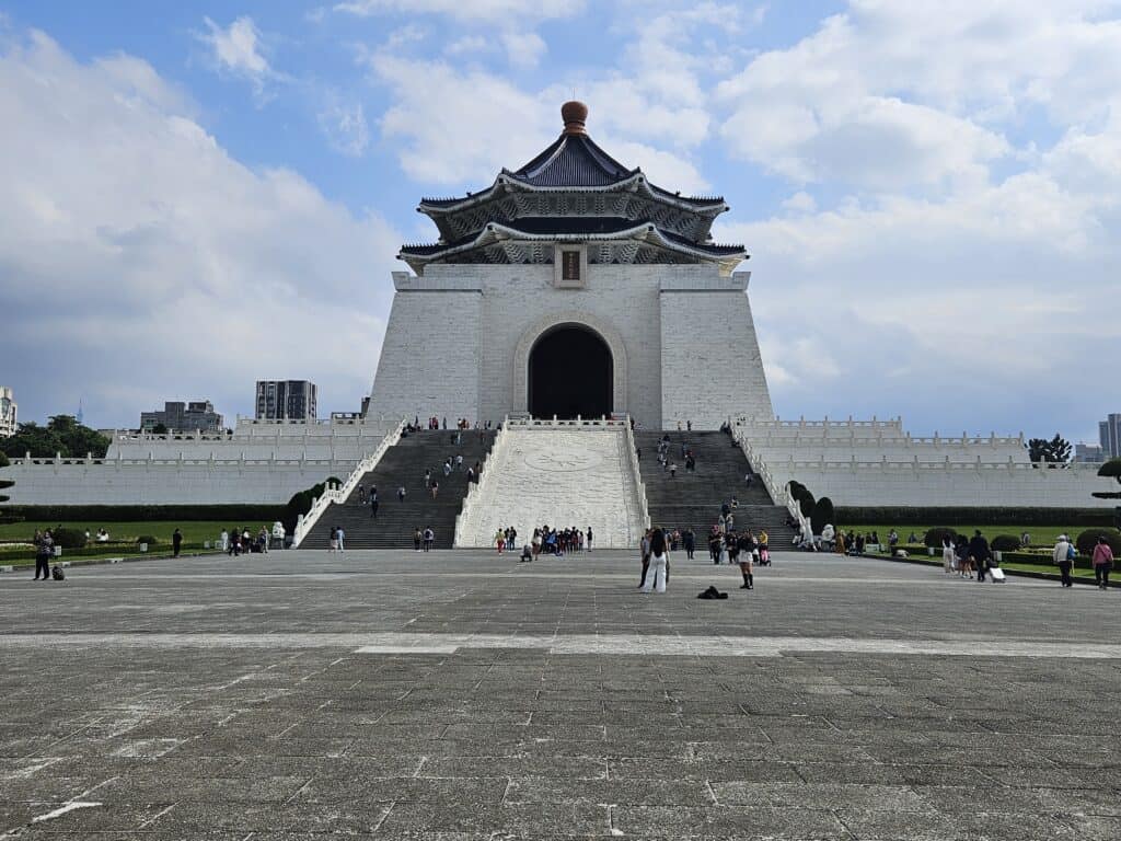 The Chiang Kai-shek Memorial Hall in Taipei, a grand white building with a blue octagonal roof and a wide staircase leading up to its arched entrance. Visitors are scattered across the expansive plaza under a partly cloudy sky.
