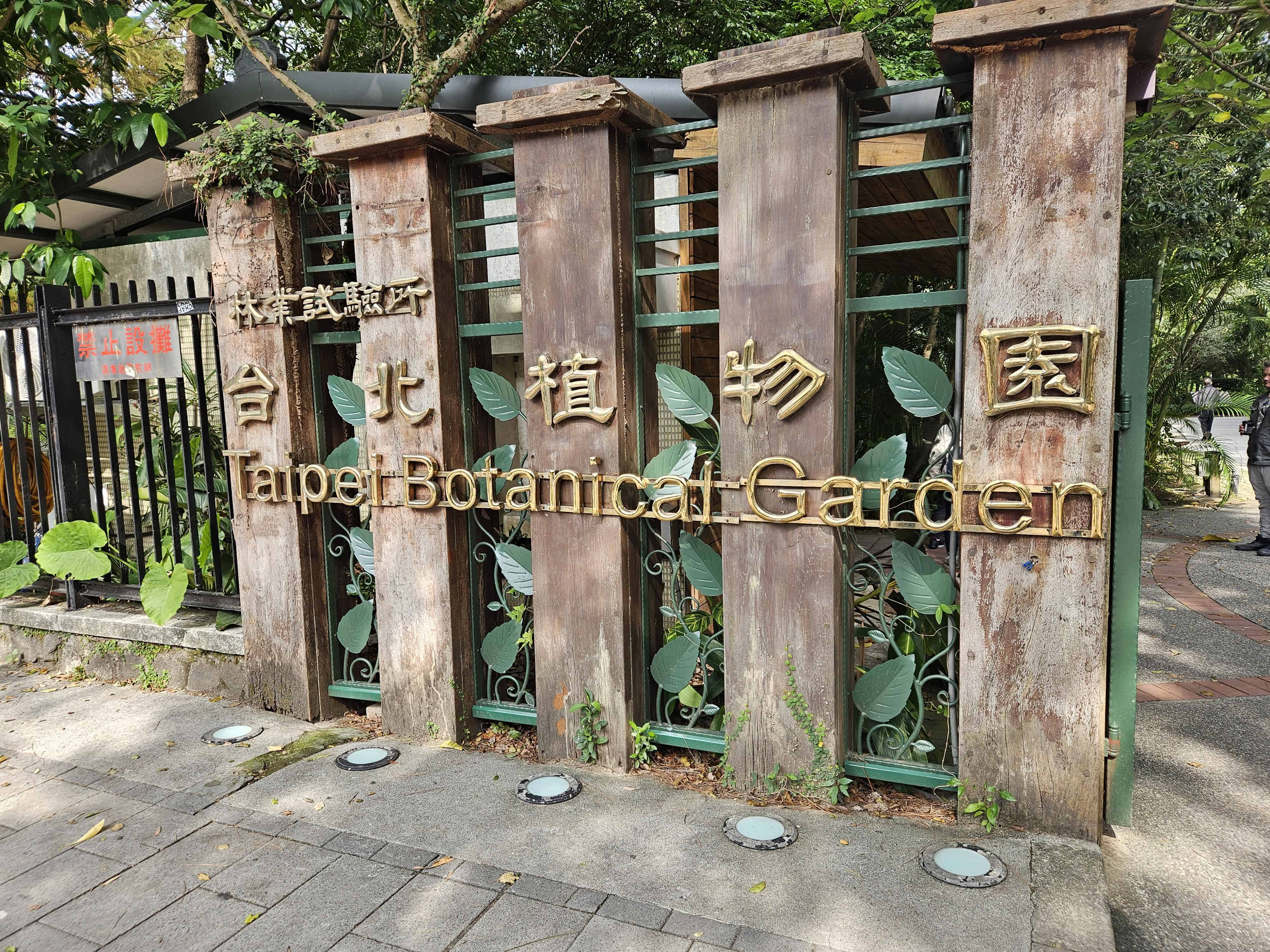 Entrance sign for the Taipei Botanical Garden, featuring gold lettering in both Chinese and English mounted on tall stone pillars with green foliage behind a metal fence. The sidewalk in front is dappled with sunlight.