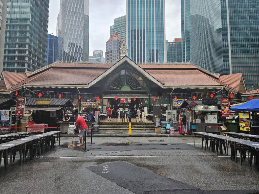 A rainy day scene at Singapore’s Satay Street, with a man mopping the wet ground in front of food stalls and long communal tables. The colonial-style market building is surrounded by modern skyscrapers, blending heritage architecture with the city’s urban skyline.