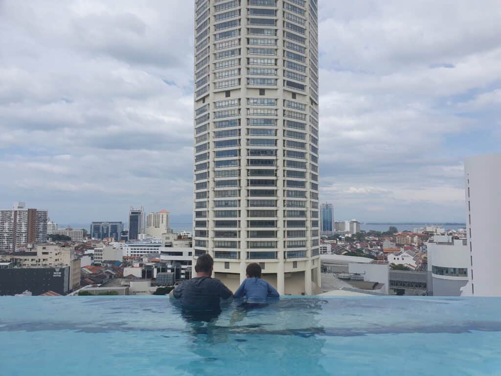 Two guests relaxing at the infinity pool of The Granite Luxury Hotel in Penang, overlooking the George Town skyline with the iconic KOMTAR tower in the background.