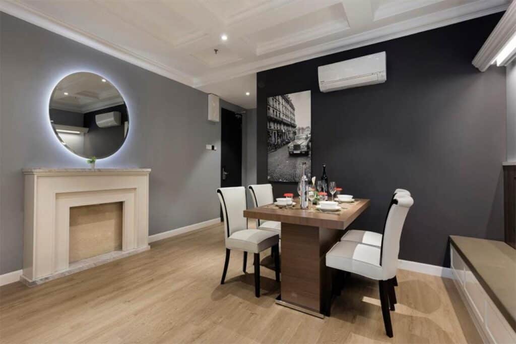 The dining area of The Razzle Two-Bedroom Suite at The Granite Luxury Hotel Penang, featuring a modern wooden table set for four, elegant white chairs, a round illuminated mirror above a decorative mantel, and stylish dark gray walls.