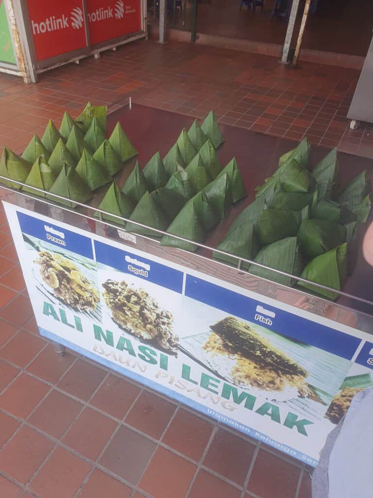 A street food stall in Penang selling “Ali Nasi Lemak,” with dozens of green banana-leaf-wrapped rice packets neatly stacked in rows. The sign shows pictures of nasi lemak served with prawns, squid, or fish, highlighting this classic Malaysian dish wrapped in daun pisang (banana leaf).