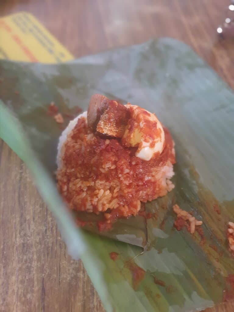 Close-up of a serving of nasi lemak unwrapped from its banana leaf, showing rice topped with spicy sambal, a piece of egg, and what looks like prawn or fish in a rich red sauce. The vibrant colors and leaf presentation highlight this traditional Malaysian dish enjoyed in Penang.
