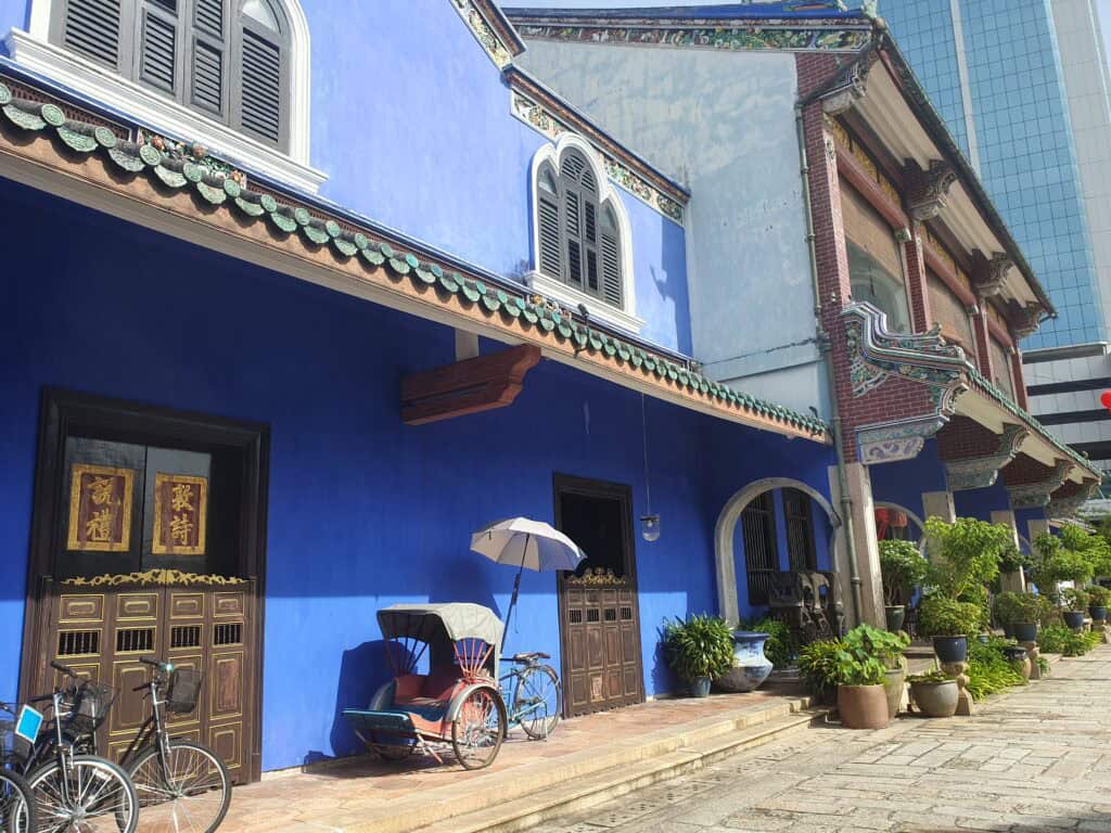 A bright blue heritage mansion in Penang, known as the Cheong Fatt Tze Mansion or Blue Mansion, with ornate wooden doors, traditional rickshaws, bicycles, and potted plants along the entrance. The vibrant colors and intricate details showcase its historical and cultural significance.