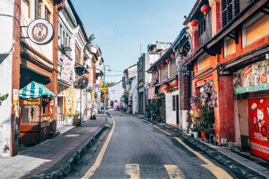 Colorful street scene in George Town, Penang, with heritage shophouses, hanging red lanterns, signs, and small shops lining a narrow road under a blue sky.