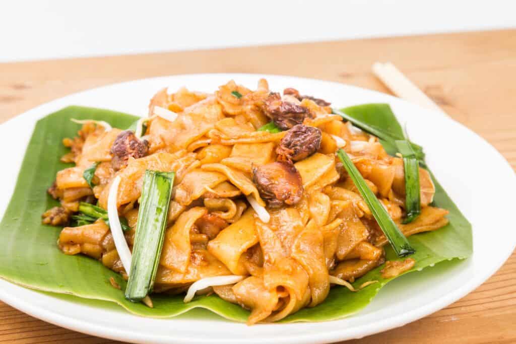 Plate of char kway teow, a stir-fried flat noodle dish with clams, bean sprouts, and green onions, served on a banana leaf.