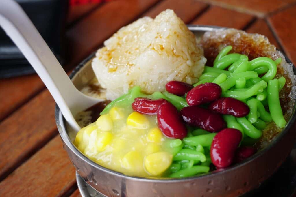 Bowl of cendol, a Malaysian dessert with shaved ice, green pandan noodles, red beans, corn, sticky rice, and sweet syrup.