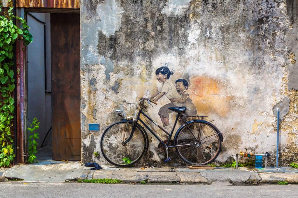 A famous street art mural in George Town, Penang, showing two children joyfully riding on a bicycle, where the painted figures blend with a real bicycle mounted against a weathered wall.