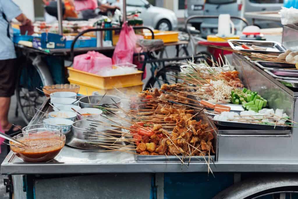 A vibrant street food stall in Penang, Malaysia, displaying skewers of meat, seafood, vegetables, and tofu ready to be cooked, with bowls of sauces and condiments on the side.