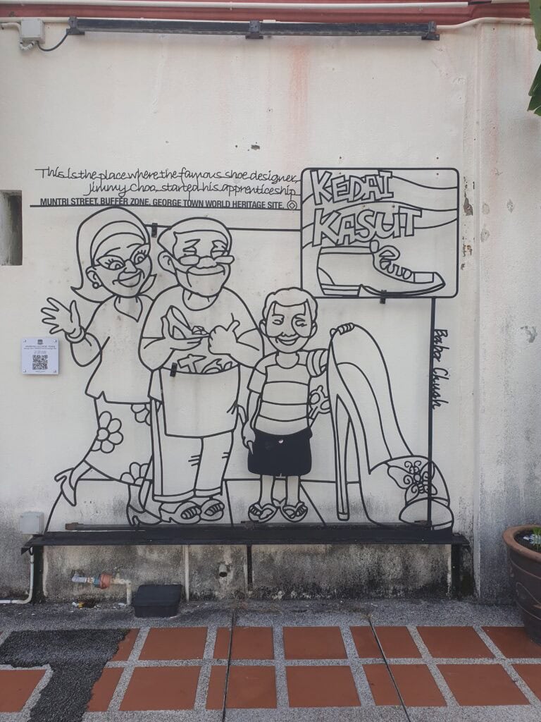 A wire art mural on a wall in George Town, Penang, depicting cartoon figures outside a shoe shop with the words “Kedai Kasut” and a high heel, marking the place where famous shoe designer Jimmy Choo started his apprenticeship.