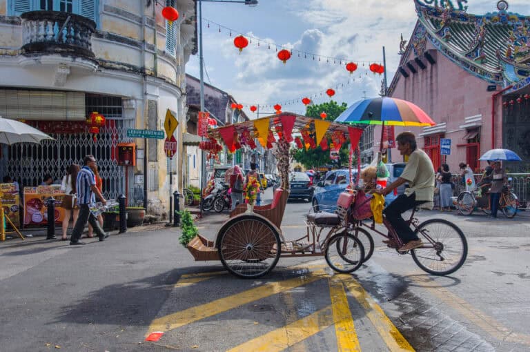 A colorful trishaw decorated with flowers and a rainbow umbrella rides through a lively street in Penang, Malaysia, with lanterns and bunting hanging above and people walking past historic shophouses and a temple.