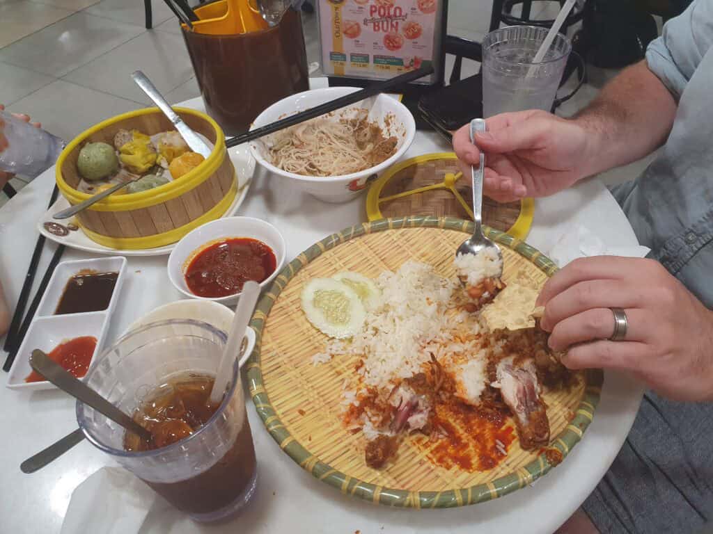 A table full of Penang food, featuring a bamboo steamer of colorful dim sum, a plate of nasi lemak with fried chicken, sambal, rice, peanuts, and cucumber, plus a bowl of noodles and sauces on the side. Iced drinks and dipping sauces complete the lively dining scene.