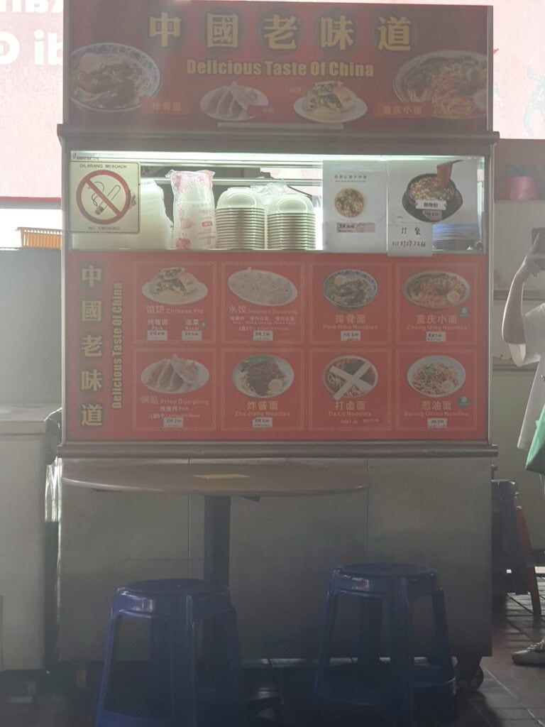 A hawker stall in Penang with a bright red sign reading “Delicious Taste of China,” offering a variety of Chinese dishes. The menu displays items like steamed and fried dumplings, pork rib noodles, Zha Jiang noodles, and spring onion noodles, with bowls and utensils stacked neatly above the counter.
