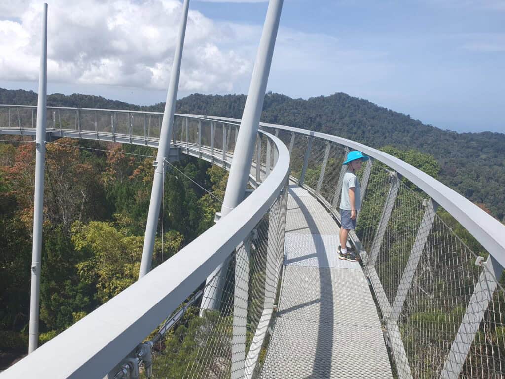 A child wearing a bright blue hat stands on the curved canopy walk at The Habitat on Penang Hill, with views of the lush forest and mountains around.