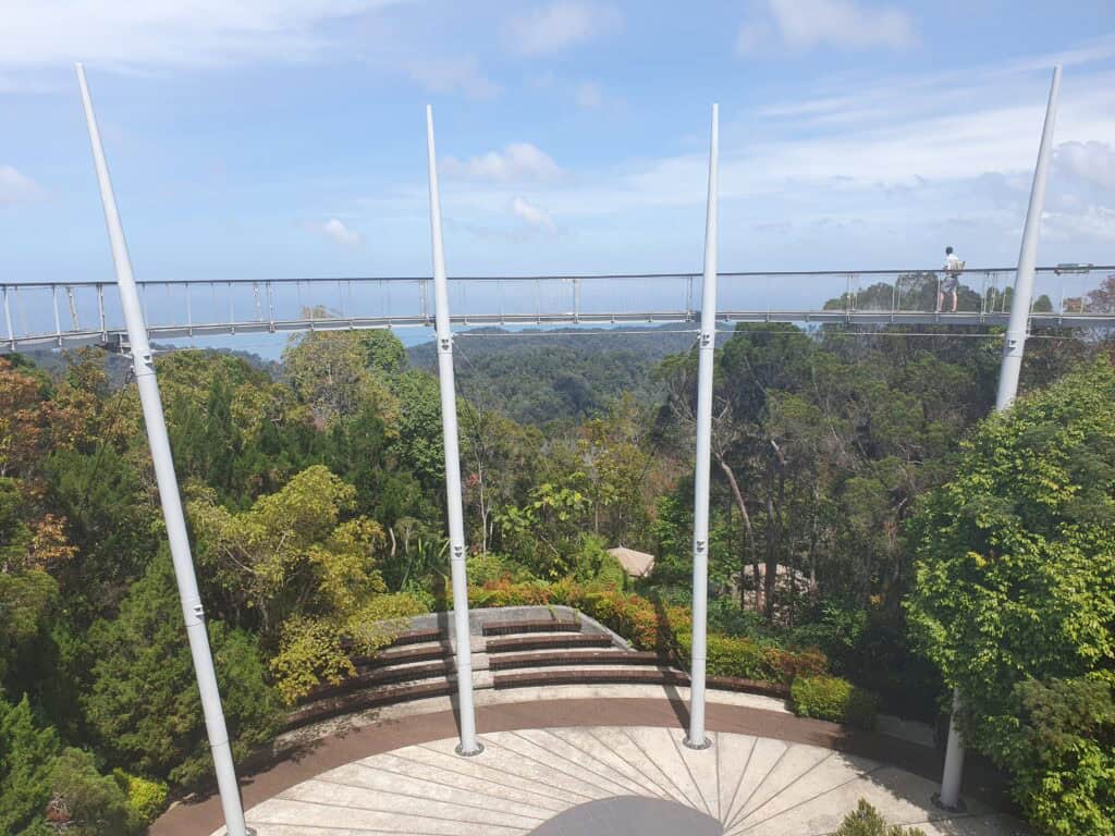 The canopy walk at The Habitat on Penang Hill, stretching above the trees with a circular platform below and green forested hills in the background.