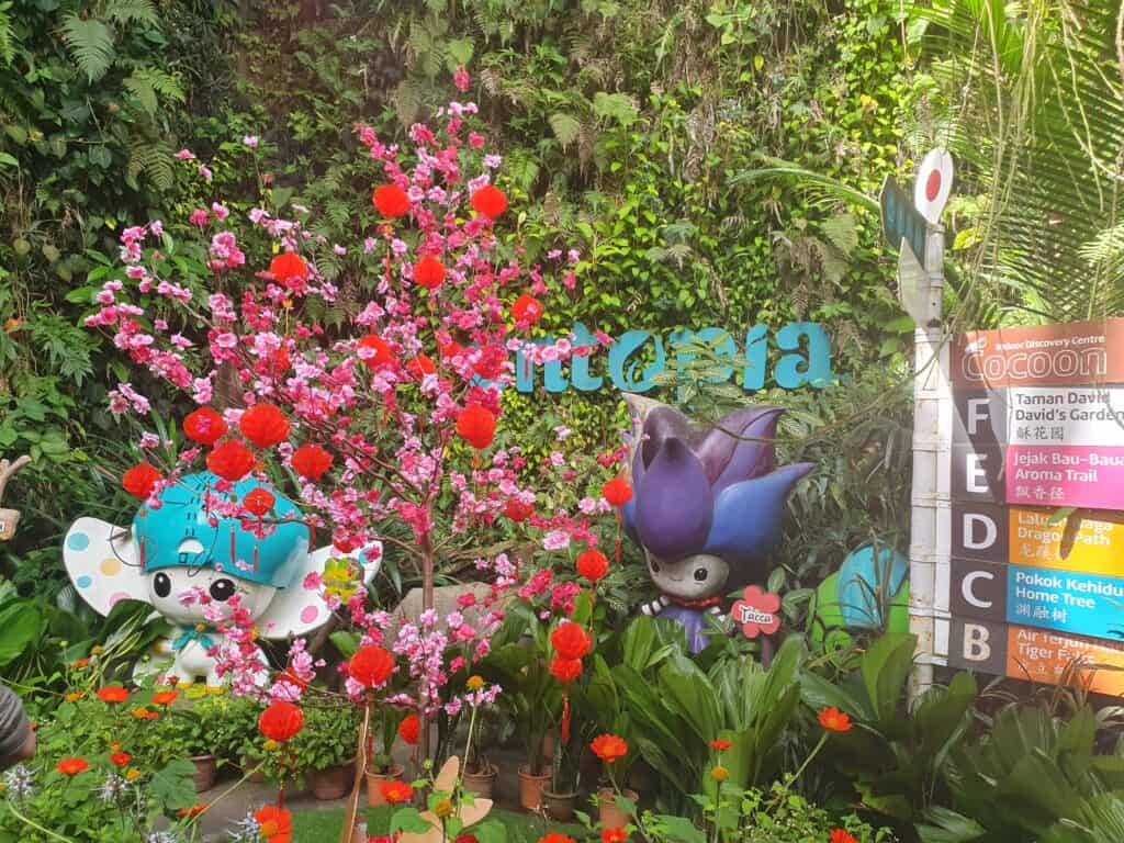 A colorful garden display at Entopia in Penang featuring playful character statues, bright pink and red blossoms, and a signpost pointing to various themed areas like David’s Garden, Aroma Trail, and Dragon Path.