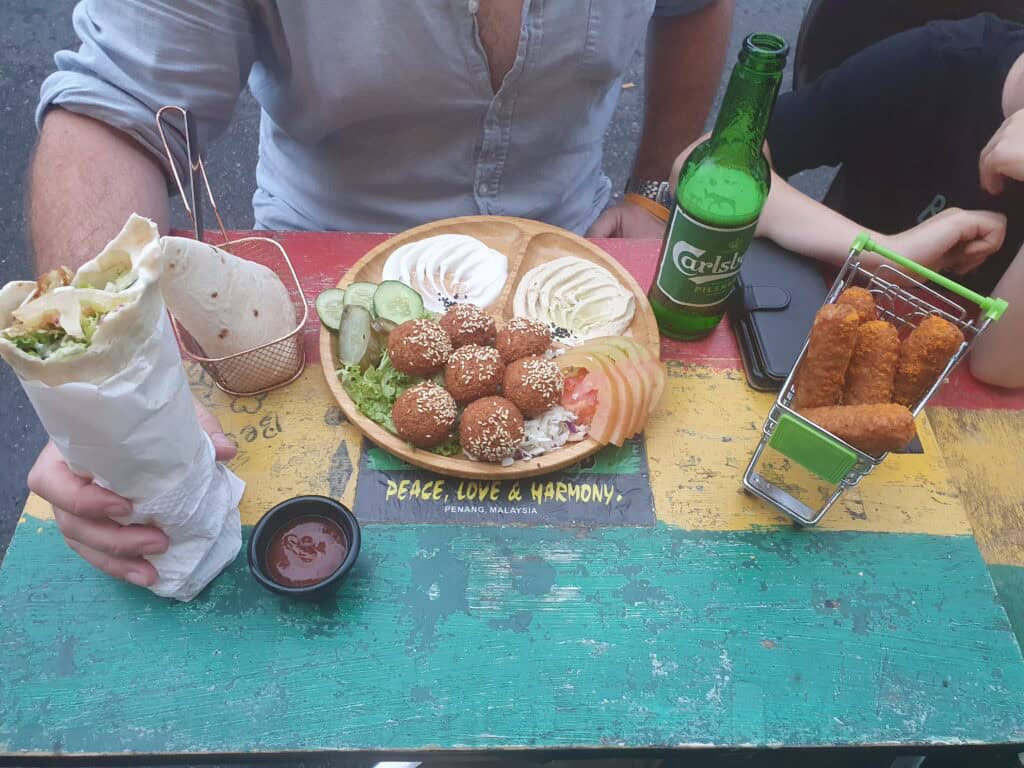 A colorful table at a Penang eatery with a vibrant “Peace, Love & Harmony” sign, serving Middle Eastern-style food. The spread includes a falafel platter with hummus, salad, pita, and sesame falafel, a wrap, a small basket of fried sticks, and a Carlsberg beer bottle.