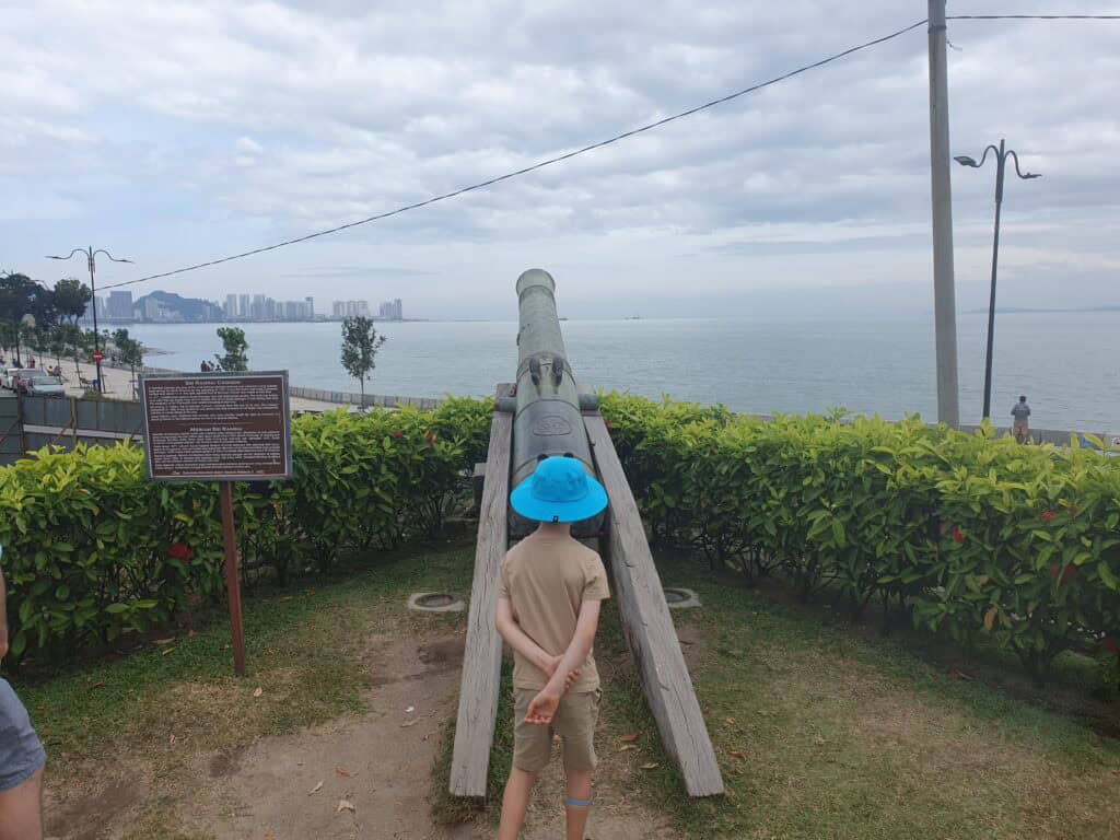 A child in a blue hat standing in front of the historic Seri Rambai Cannon on the seafront in Penang, with a view of the ocean and city skyline in the distance.