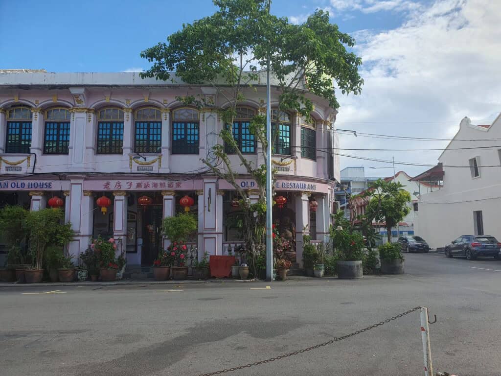 A pastel pink heritage shophouse in Penang, Malaysia, housing the Old House Restaurant, with red lanterns, potted plants, and traditional Chinese architectural details under a blue sky.