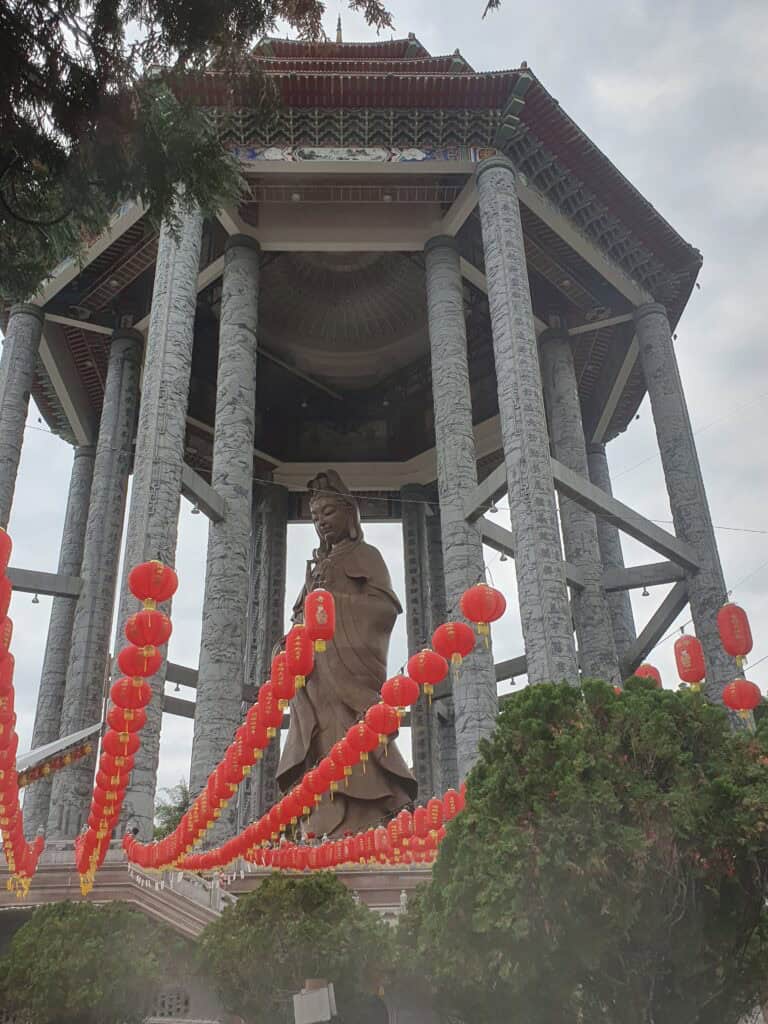 Wide view of the towering Kuan Yin statue standing gracefully under a grand pavilion, with strings of bright red lanterns and greenery in the foreground.