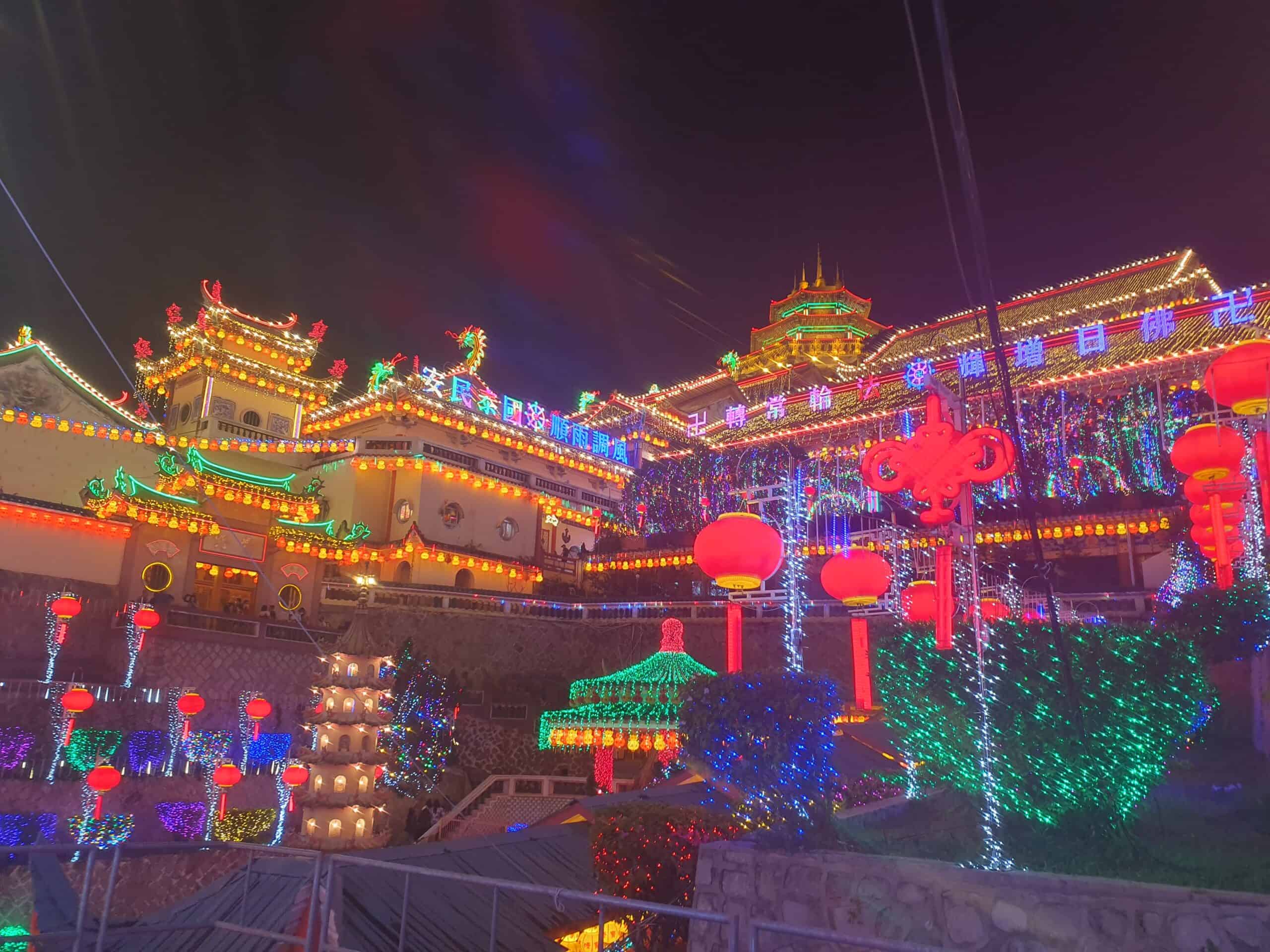 A brightly illuminated Kek Lok Si temple in Penang at night, covered in colorful lights, lanterns, and glowing decorations, creating a festive and vibrant scene. The building features traditional Chinese architectural details and intricate rooftop designs.