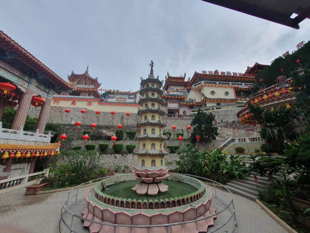 Multi-tiered pagoda with a lotus-shaped fountain at its base, surrounded by red lanterns and colorful temple buildings of Kek Lok Si Temple in Penang, Malaysia, set against a cloudy sky.