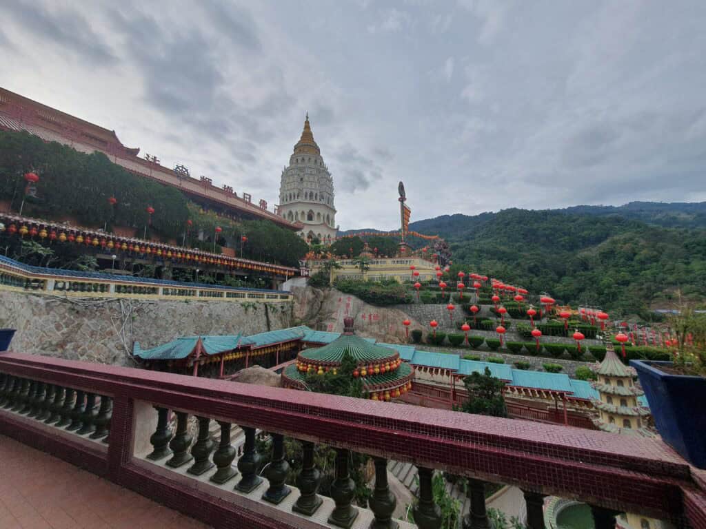 Daytime view of Kek Lok Si Temple complex, with its iconic white and gold pagoda surrounded by lush green hills and red lanterns lining the terraces.
