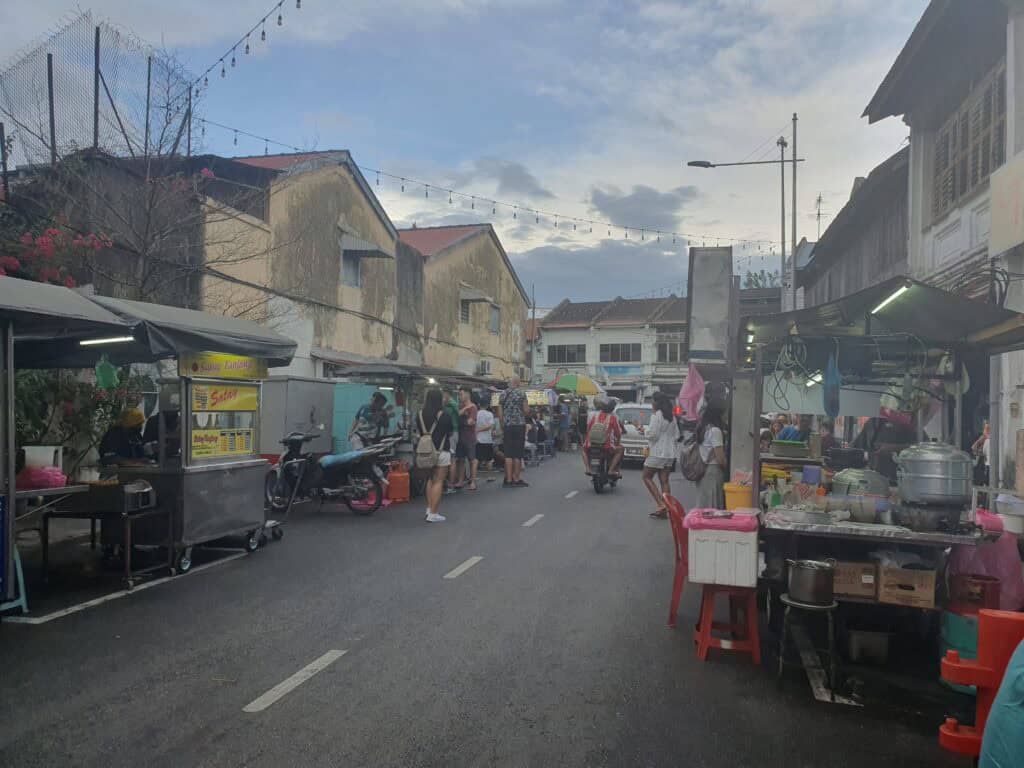 A bustling evening street food market in Penang with food stalls lining both sides of the narrow street. People are queuing and mingling, with vendors preparing dishes under canopies and strings of lights hanging overhead, creating a lively and inviting atmosphere.