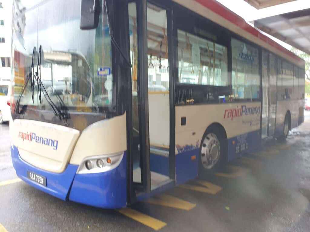 A Rapid Penang public bus parked at a covered stop on a rainy day, with its door open and wet pavement visible around it.