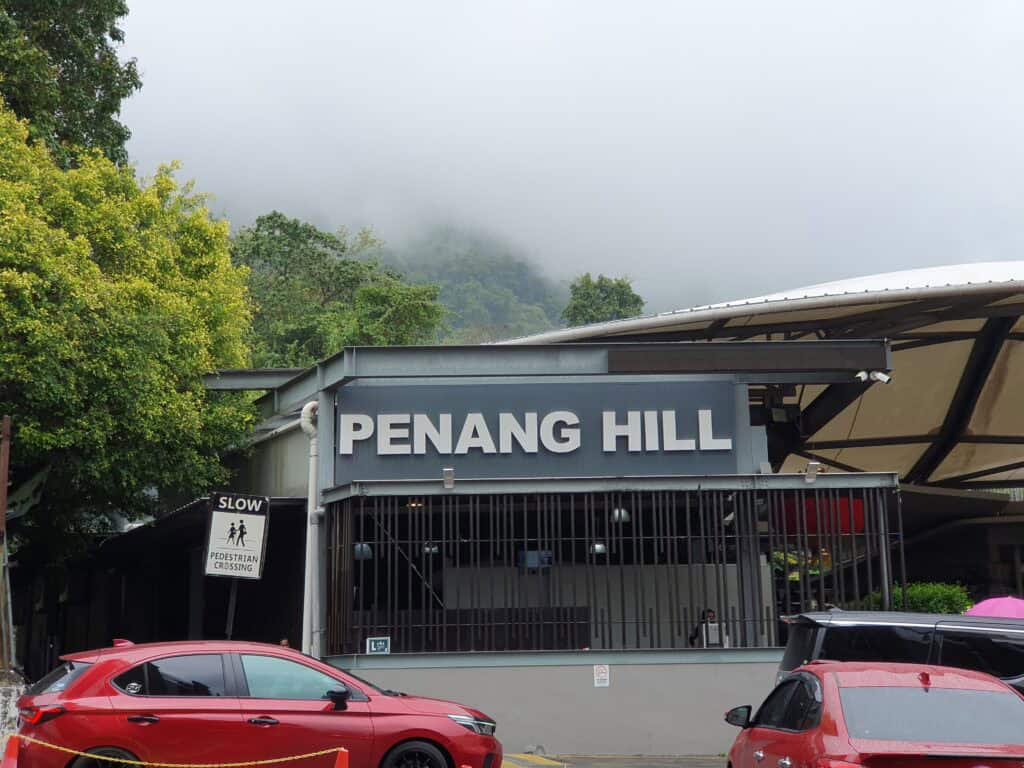 The entrance to Penang Hill station on a misty day, with the name displayed in large letters and cars parked in front, a popular family attraction in Penang.