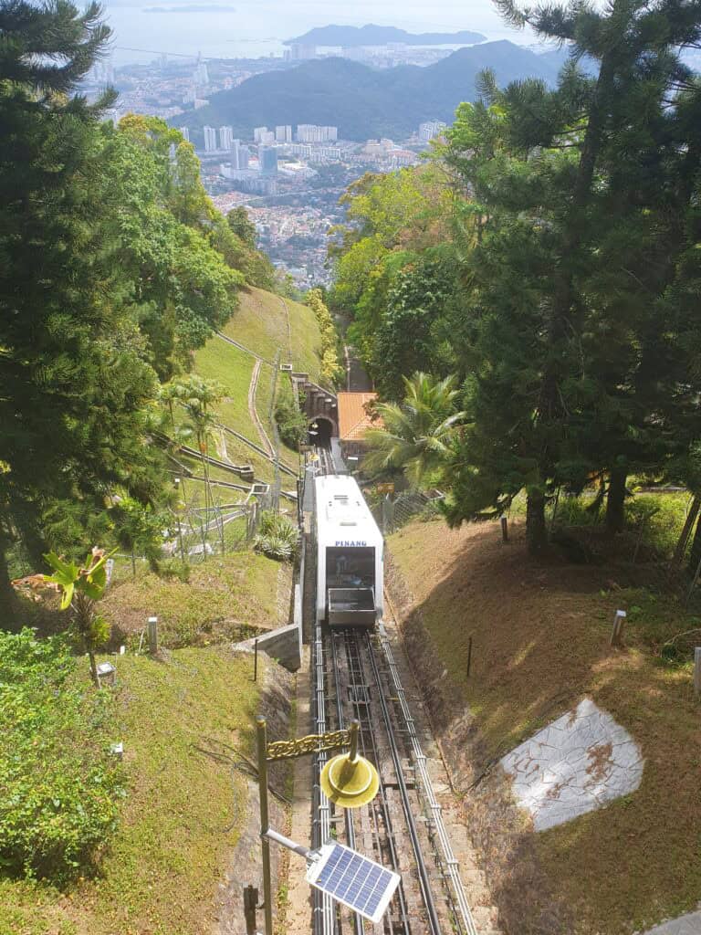 The Penang Hill funicular train making its way up the lush green hillside, with panoramic views of George Town and the sea in the distance — a fun and scenic ride for families with kids.