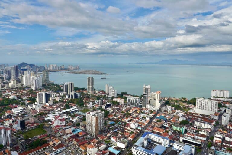 A panoramic aerial view of George Town, Penang, Malaysia, showing a mix of high-rise buildings, colorful shophouses, and the coastline with the sea and distant mountains under a partly cloudy sky.