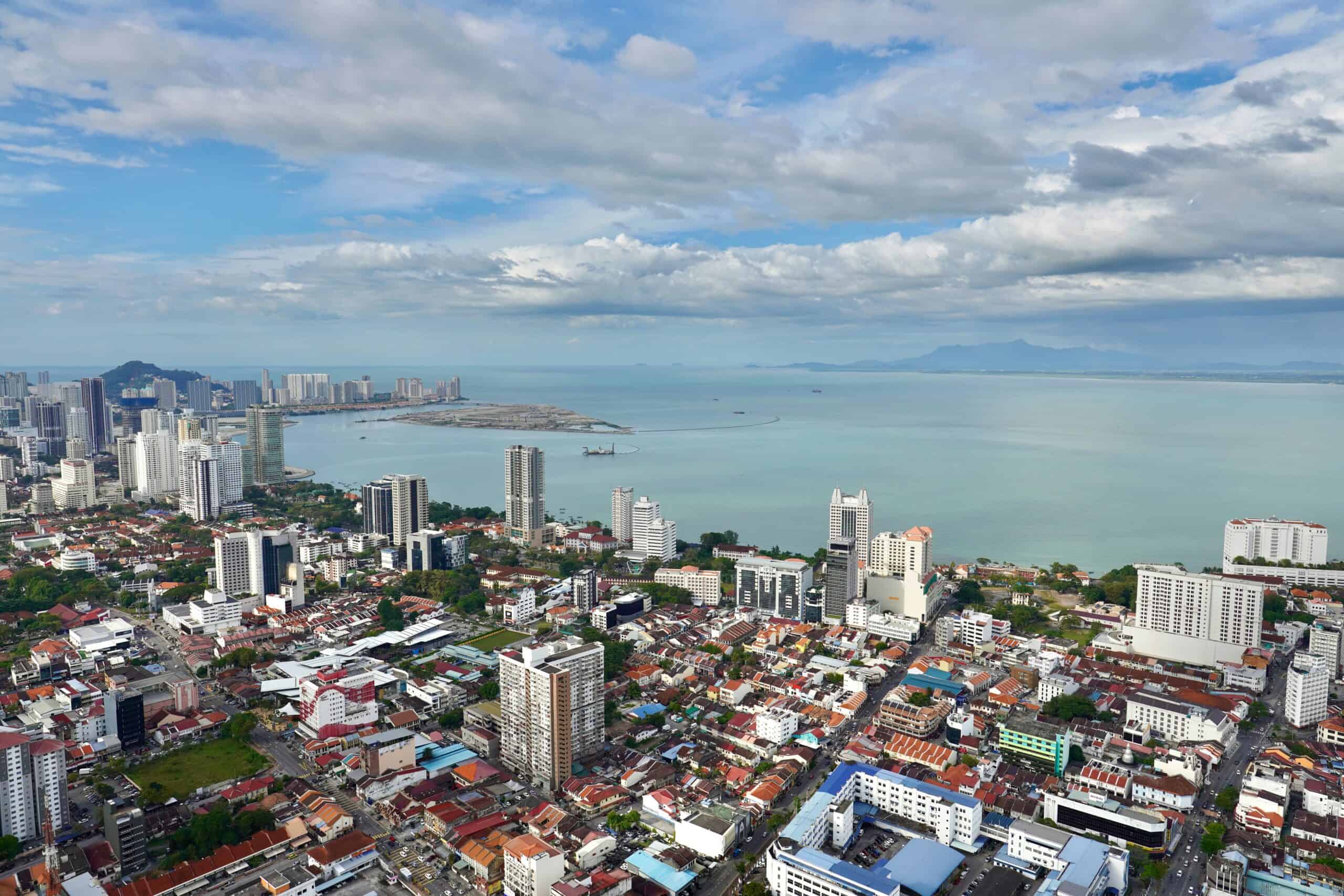 A panoramic aerial view of George Town, Penang, Malaysia, showing a mix of high-rise buildings, colorful shophouses, and the coastline with the sea and distant mountains under a partly cloudy sky.
