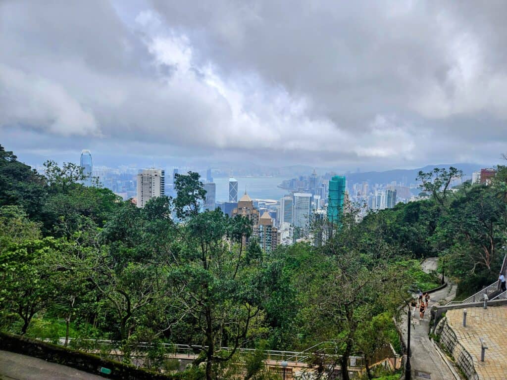 A sweeping vista from a hillside trail showing dense green treetops, a winding path below, and the high-rise buildings of Hong Kong stretching to the harbor in the distance.