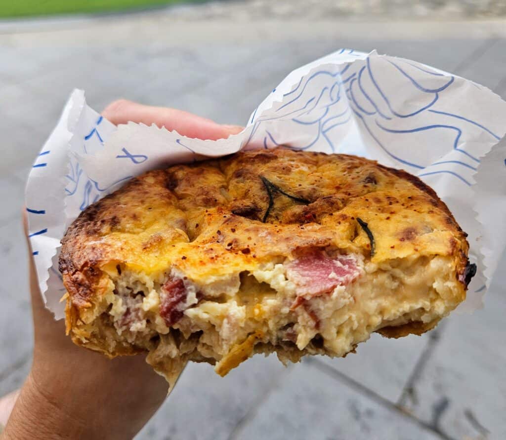 A close-up of a hand holding a partially eaten savory pastry filled with creamy egg, ham, and cheese, wrapped in branded white paper with blue line art.