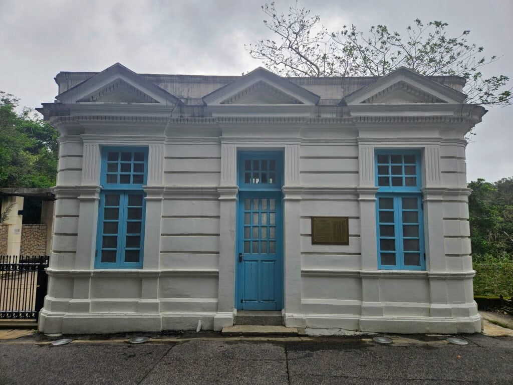 A neoclassical-style white building with blue doors and window frames, featuring columns and triangular pediments, with a brass plaque mounted beside the entrance.