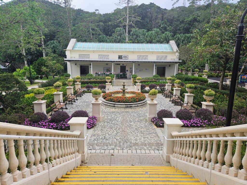 A manicured public garden viewed from a staircase, featuring symmetrical flower beds, ornamental planters, benches, and a central fountain, set against a backdrop of lush forest and a public building.