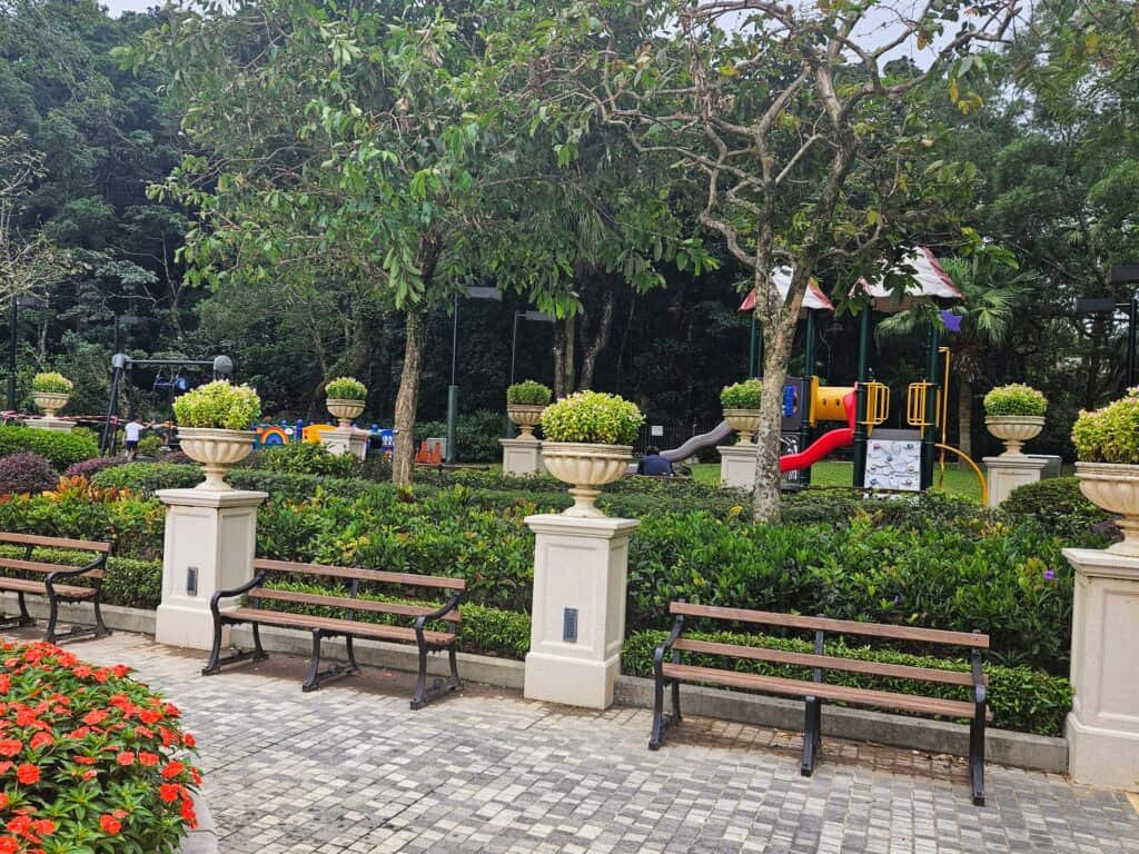 A children’s playground with colorful slides and climbing equipment set in a landscaped park, surrounded by trimmed hedges, benches, and decorative planters.