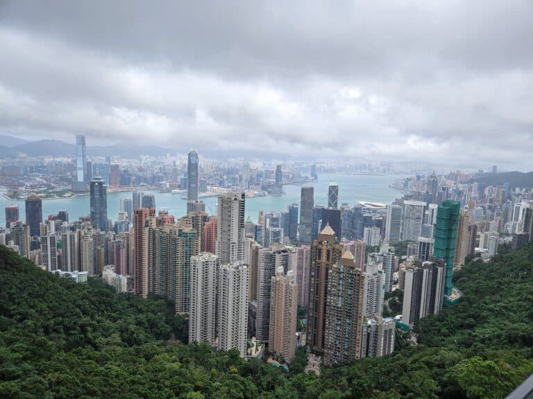 A panoramic view of Hong Kong’s skyline with densely packed skyscrapers, Victoria Harbour, and distant mountains under thick gray clouds, seen from a high vantage point.