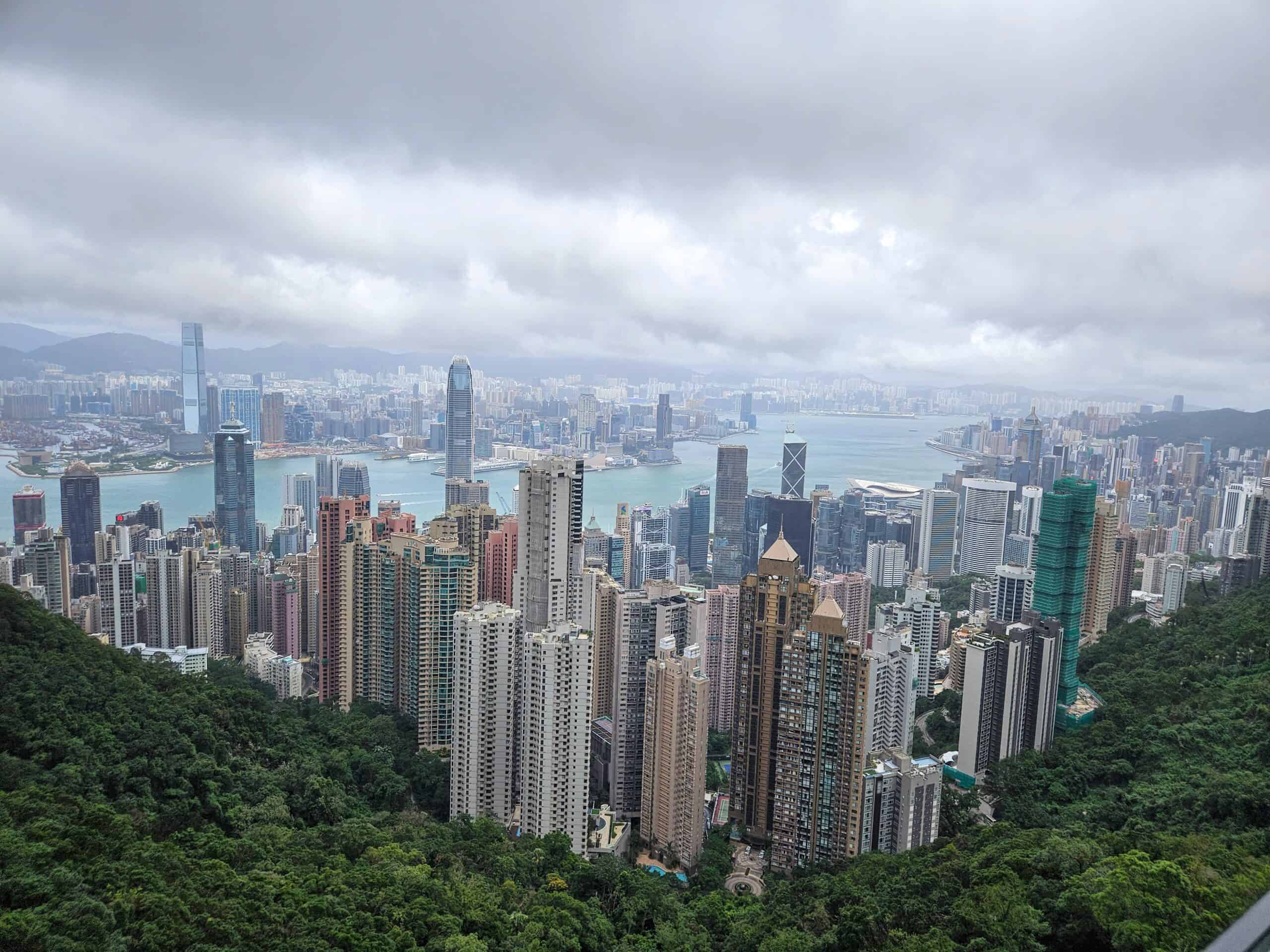 A panoramic view of Hong Kong’s skyline with densely packed skyscrapers, Victoria Harbour, and distant mountains under thick gray clouds, seen from a high vantage point.