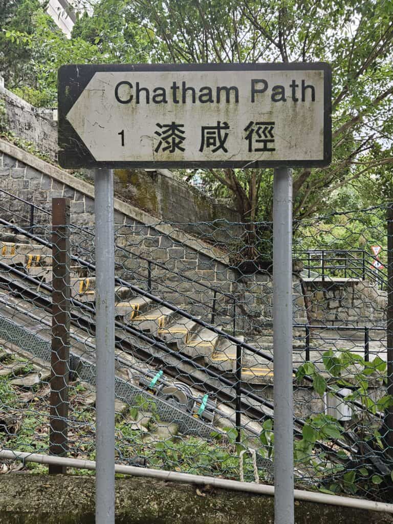 A weathered directional sign for Chatham Path with English and Chinese text, mounted on two poles near a stone staircase and chain-link fence, surrounded by lush greenery.