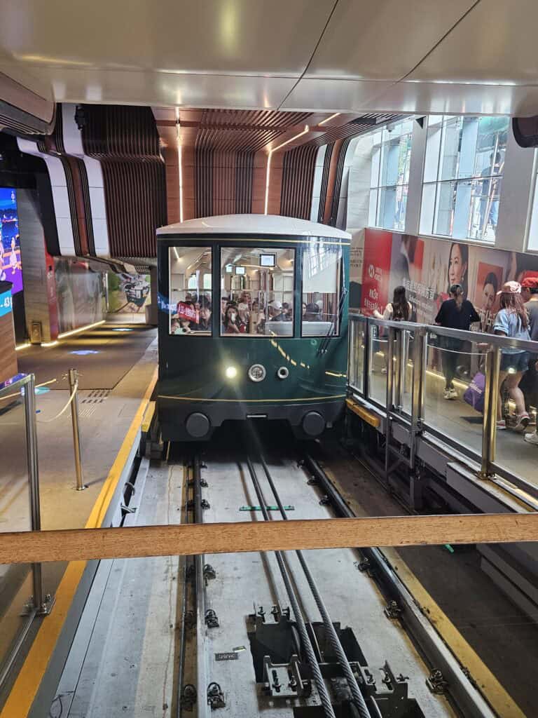 A green Peak Tram car at the station platform with passengers inside, surrounded by modern interior architecture and tourists waiting along the boarding area.