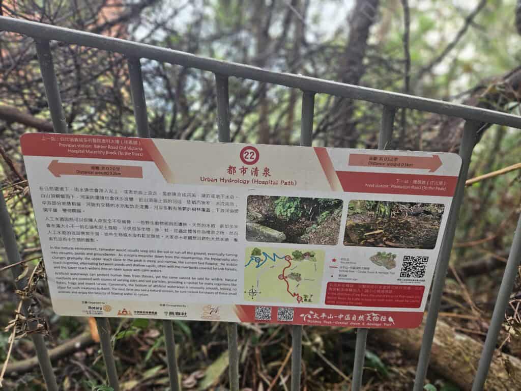 An educational sign titled "Urban Hydrology (Hospital Path)" displayed on a metal railing, explaining water flow in nature and its impact on wildlife, with supporting photos, a map, and bilingual text.