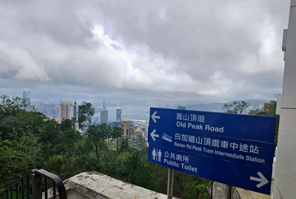 A panoramic view of Hong Kong's skyline under dramatic clouds, with a blue directional sign in the foreground pointing to Old Peak Road, Barker Road Peak Tram station, and public toilets.