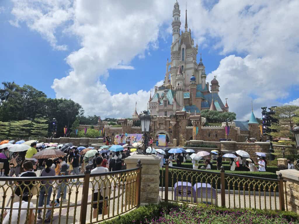 A crowd of visitors with umbrellas gathers in front of the towering Castle of Magical Dreams at Hong Kong Disneyland under a bright blue sky. The whimsical castle, with its colorful turrets and ornate design, stands as the park’s fairytale centerpiece.