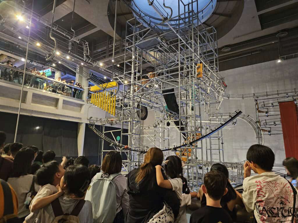 A crowd of children and adults watches a towering kinetic sculpture made of metal tracks and structures inside the Hong Kong Science Museum. The complex installation features rolling balls, loops, and mechanical elements that demonstrate motion and physics in action.