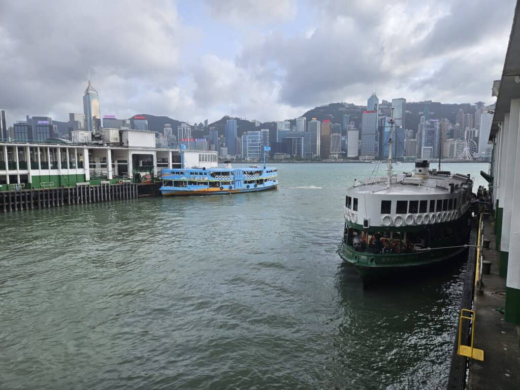 Two Star Ferry boats docked at the terminal in Victoria Harbour, with Hong Kong’s iconic skyline rising in the background under a cloudy sky. The green and white ferries contrast against the high-rise buildings and rolling hills of Hong Kong Island.