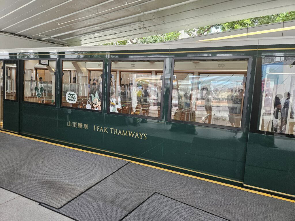 Side view of the green Peak Tram at a station in Hong Kong, with passengers visible through large windows and Disney character decals decorating the glass. The words "Peak Tramways" are printed in gold lettering on the tram’s exterior, capturing a moment before the scenic ride to Victoria Peak.