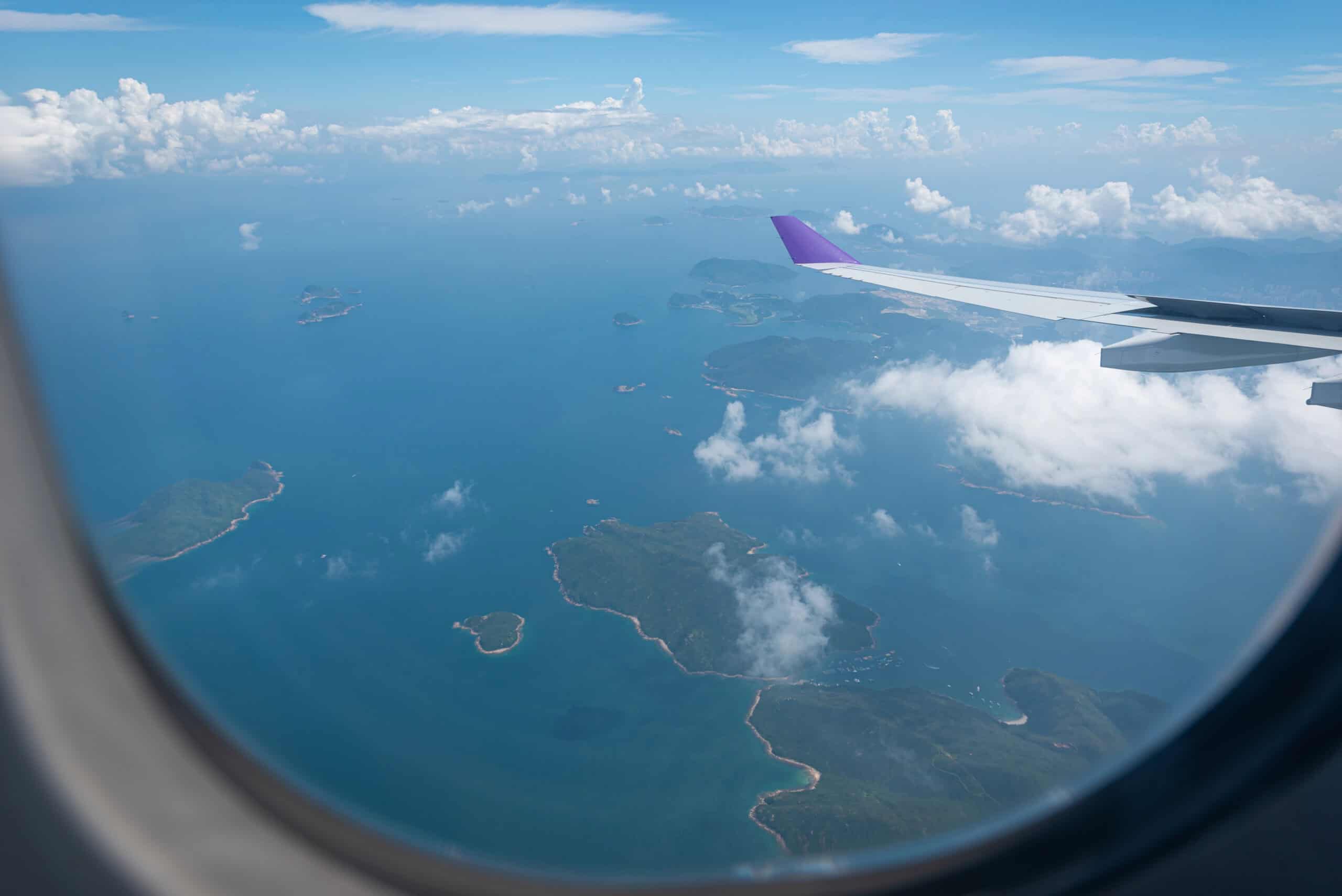 View from an airplane window showing scattered green islands surrounded by turquoise ocean under a bright blue sky with fluffy clouds. The aircraft wing with a purple tip is visible, capturing the feeling of flying between destinations on a multi-city trip.